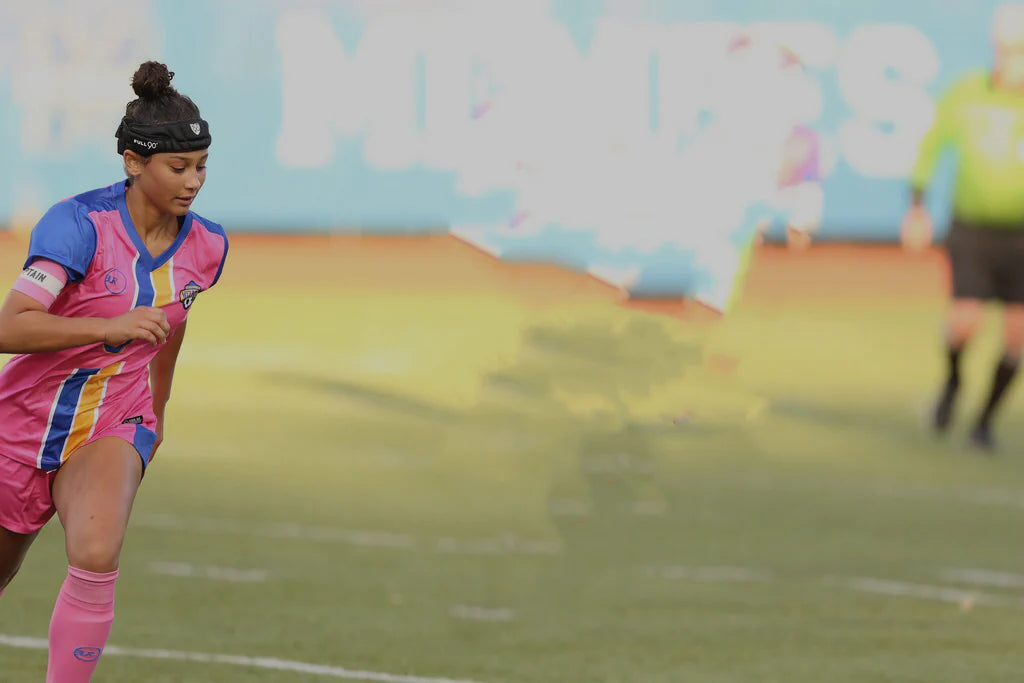 Female soccer player wearing a pink and blue uniform with a captain's armband, running on a field during a match.